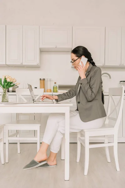 Боковой вид бизнесвумен в блейзере над пижамой, работающей на кухне, говорящей по смартфону и использующей ноутбук — стоковое фото