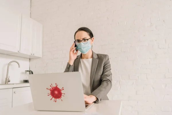 Serious businesswoman in medical mask and eyeglasses talking on smartphone near laptop with coronavirus bacteria sticker — Stock Photo
