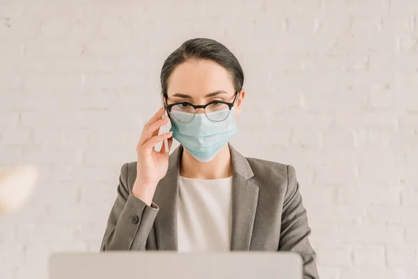 Giovane donna d'affari in maschera medica e occhiali che parla su smartphone a casa — Foto stock