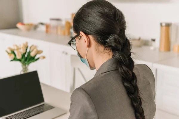 Брюнетка, длинноволосая деловая женщина в медицинской маске работает на ноутбуке на кухне — стоковое фото