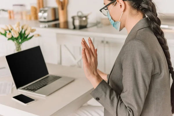 Junge Geschäftsfrau in medizinischer Maske zeigt Hallo-Geste bei Videochat in Küche — Stockfoto