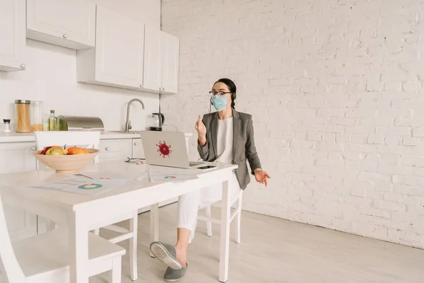 Femme d'affaires en colère dans le masque médical et blazer sur pyjama montrant majeur tout en travaillant dans la cuisine près de l'ordinateur portable — Photo de stock