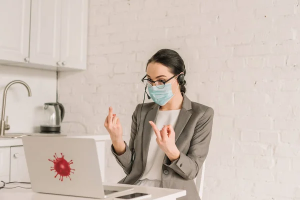 Mujer de negocios enojado en máscara médica y auriculares que muestran los dedos medios mientras trabaja en la cocina cerca de la computadora portátil - foto de stock