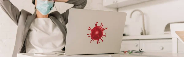 Panoramic shot of businesswoman in medical mask sitting in kitchen near laptop with coronavirus bacteria sticker — Stock Photo