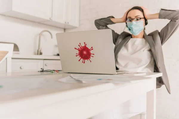 Selektiver Fokus der müden Geschäftsfrau mit medizinischer Maske und Headset, die den Kopf berührt, während sie auf Laptop mit Bakterienaufkleber in der Küche blickt — Stockfoto