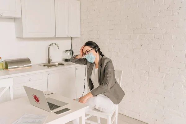 Stanca donna d'affari in giacca su pigiama, maschera medica e auricolare seduta vicino al computer portatile mentre lavora a casa — Foto stock