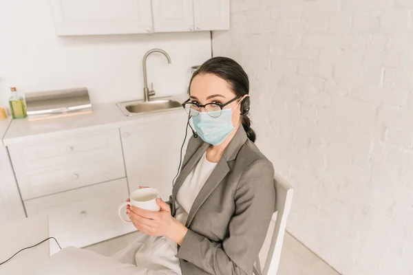 Giovane donna d'affari in maschera medica, auricolare e blazer in possesso di una tazza di caffè in cucina — Foto stock