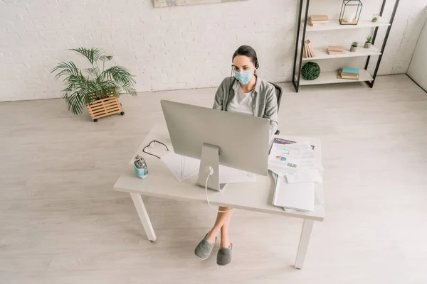 Hochwinkel-Ansicht des Fernbedieners in medizinischer Maske und Headset, der zu Hause in der Nähe des Computermonitors arbeitet — Stockfoto