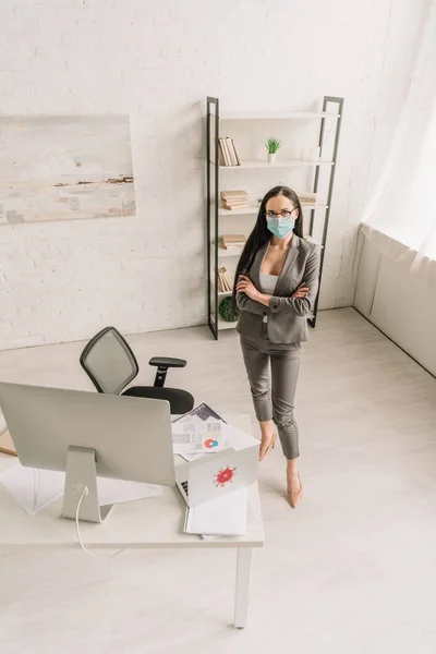 Высокий угол обзора деловой женщины в формальной одежде и медицинской маске, стоящей со скрещенными руками возле стола с компьютерами дома — стоковое фото