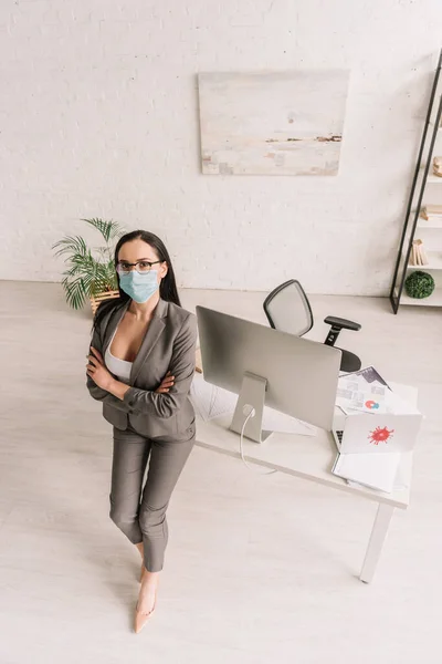 High angle view of businesswoman in formal wear and medical mask standing with crossed arms near table with computers at home — Stock Photo