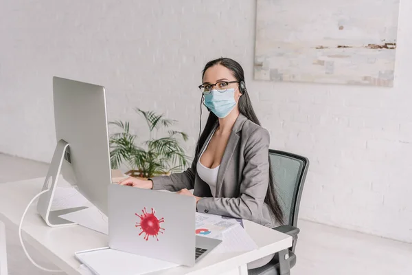 Geschäftsfrau mit medizinischer Maske und Kopfhörer blickt in die Kamera, während sie zu Hause in der Nähe von Laptop und Computermonitor arbeitet — Stockfoto