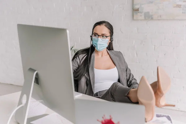 Selective focus of businesswoman in medical mask and headset working at home while holding legs on table — Stock Photo