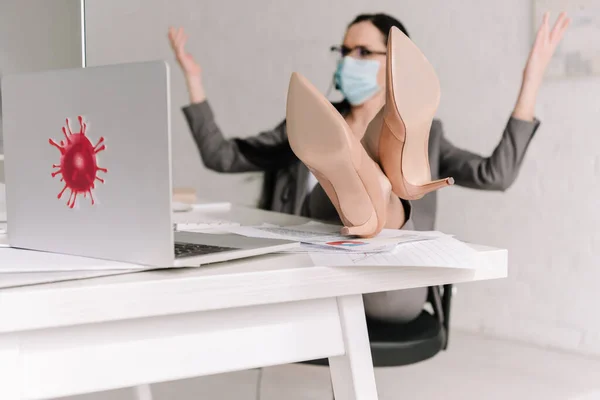 Selective focus of businesswoman in medical mask showing shrug gesture while working at home with legs on table — Stock Photo