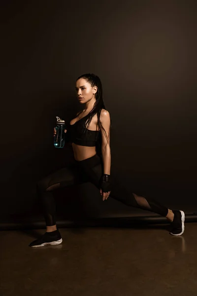 Beautiful sportswoman with sports bottle doing lunges on black — Stock Photo