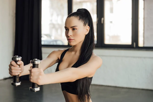Beautiful sportswoman holding dumbbells with outstretched hands in gym — Stock Photo