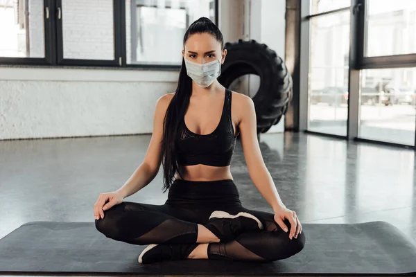 Brünette Sportlerin in medizinischer Maske sitzt mit überkreuzten Beinen auf Fitnessmatte und schaut in die Kamera im Fitnessstudio — Stockfoto
