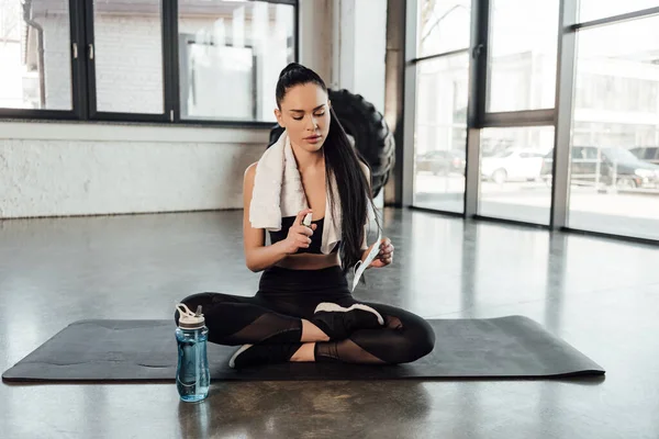 Sportswoman with crossed legs and towel using hand sanitizer and holding medical mask on fitness mat near sports bottle in gym — Stock Photo