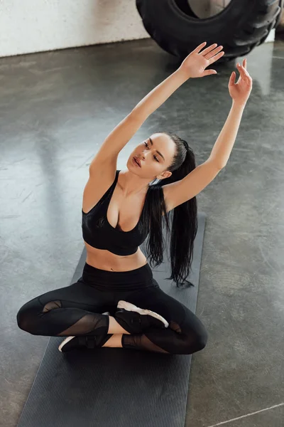 Vista de ángulo alto de la deportista con las piernas cruzadas apoyadas con las manos en el aire en la alfombra de fitness en el gimnasio - foto de stock