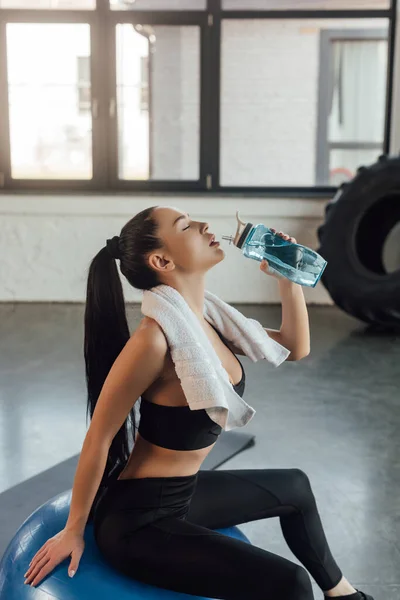 Beautiful sportswoman with closed eyes and towel drinking water and sitting on fitness ball in gym — Stock Photo