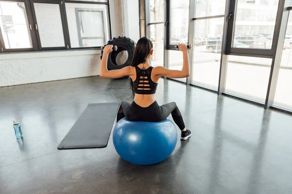 Back view of sportswoman with dumbbells on fitness ball near mat and sports bottle in gym — Stock Photo