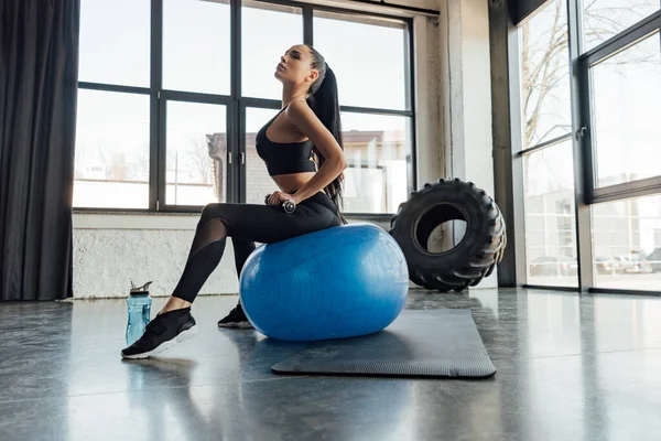 Seitenansicht einer Sportlerin mit Hantel, die auf einem Fitnessball in der Nähe einer Sportflasche im Fitnessstudio sitzt — Stockfoto