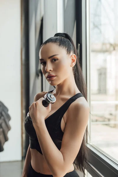 Beautiful sportswoman holding dumbbell and looking at camera near window in gym — Stock Photo
