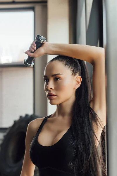 Beautiful sportswoman holding dumbbell over head in gym — Stock Photo