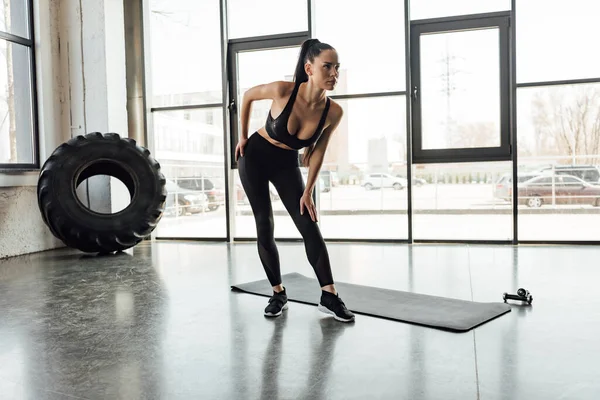 Sportiva bruna appoggiata vicino al tappeto fitness e ai manubri in palestra — Foto stock