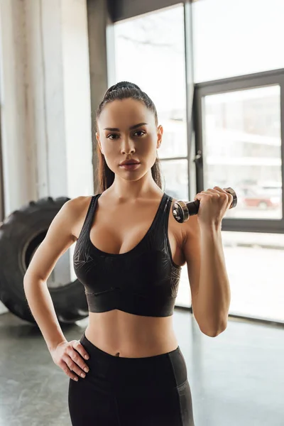 Deportista morena con la mano en el entrenamiento de cadera con mancuerna y mirando a la cámara en el gimnasio - foto de stock