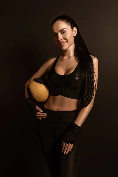 Morena deportista con bola mirando a la cámara y sonriendo aislado en negro - foto de stock