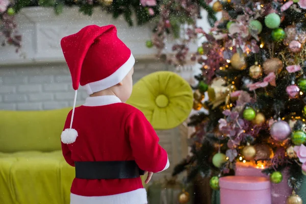 Noel ağacının yanında oturan takım elbiseli çocuk hediyelerin getirilmesini bekliyor.
