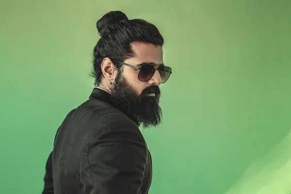 side view of handsome bearded man from the back with a man bun hairstyle posing in studio wearing a suit and sunglasses on green background, male model isolated fashion portrait photograph.