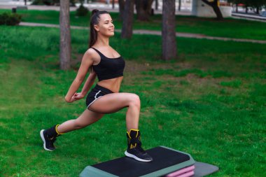 Genç, hoş bir kadın parkta adım platformunda egzersiz yapıyor.