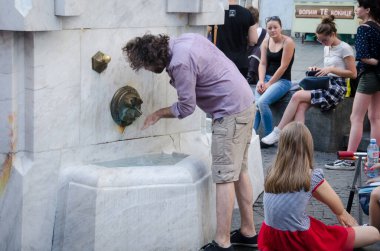 Uzun saçlı bir adam, Sırbistan 'ın Belgrad kentinin yaya caddesinde sıcak bir günde çeşmeden su içiyor