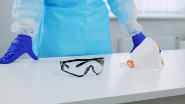 Close-up of a glasses and respirator on a white table in hospital. On the background a doctor in protective gloves and suit leaning hands on a table. The camera slowly moves to the side — Stock Video