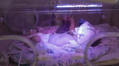 Bebek kuvözde yatıyor. Beslenme tüpü takılı. Bebek bacaklarını hareket ettiriyor, yandan bakıyor. Yoğun bakım, yeni doğan doktorlar gözetiminde..