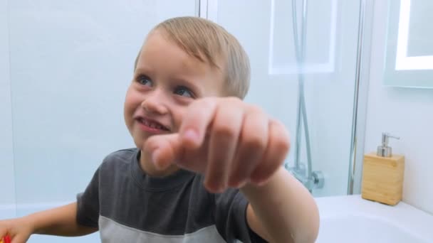 Sonriente niño alegre en una camiseta gris con un cepillo de dientes en las manos señalando con el dedo al espectador mirando en la pantalla y mostrando el gesto en el baño blanco claro — Vídeos de Stock