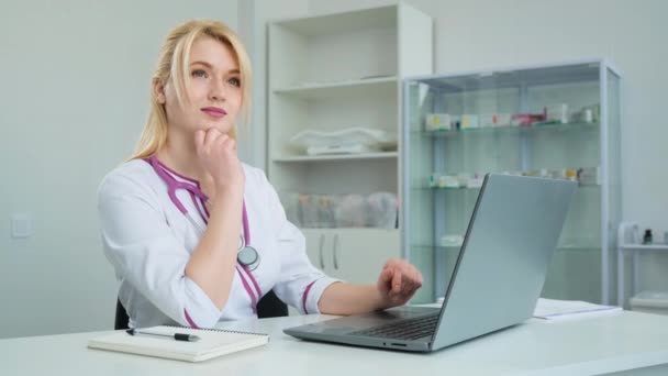 Jonge blanke vrouw arts in witte jas zit aan tafel in het kabinet. Vrouwelijke arts op zoek naar laptop computer en rekening houdend met ziekteverschijnselen — Stockvideo