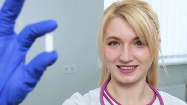 Close up de médico feminino loira caucasiana de casaco branco, estetoscópio violeta segurando uma pílula branca com os dedos em luvas azuis no consultório do hospital ou clínica. Foco muda do médico para a pílula — Vídeo de Stock