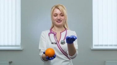 Beyaz önlüklü beyaz sarışın doktor, menekşe steteskop elinde beyaz bir hap, sağ elinde turuncu ve sol elinde mavi eldivenler ve sağlıklı bir yemeği ve gülümsemeyi seçiyor.