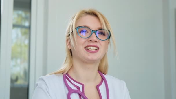 Joven doctora caucásica en bata blanca, estetoscopio y gafas mirando hacia el futuro, soñando con la futura carrera. Retrato de cerca en la clínica — Vídeos de Stock