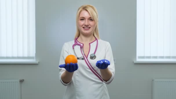 Caucasian young doctor in white medical coat, violet stethoscope holding a white pills with left hand and orange in rigth hand in blue gloves. Choice of medicine, pill or fruits and vitamins concept — Stock Video