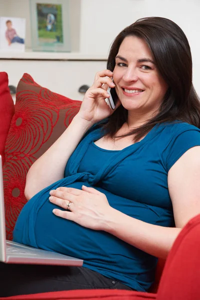 Femme enceinte assise sur le canapé en utilisant un ordinateur portable et un téléphone portable — Photo