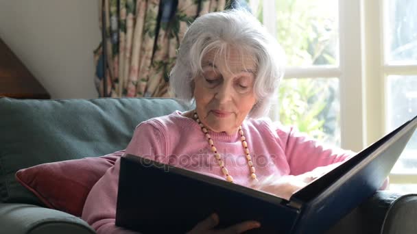 Старшая женщина в кресле дома глядя на фотоальбом — стоковое видео