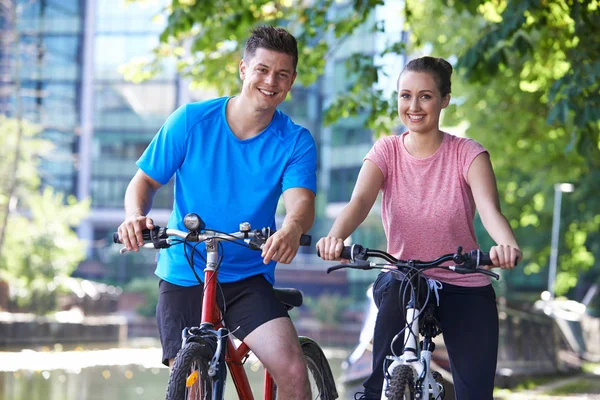 Joven pareja ciclismo junto al río en entorno urbano — Foto de Stock