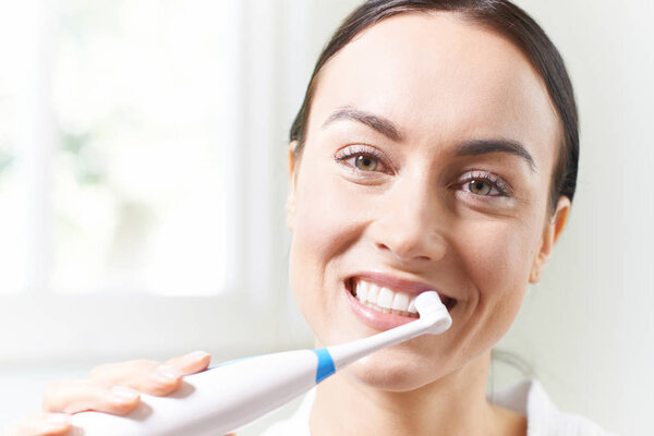 Женщина чистит зубы электрической зубной щеткой в ванной комнате
