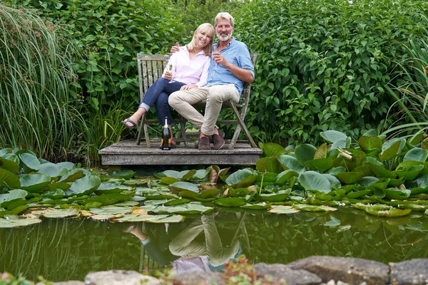 湖畔の木製桟橋の椅子に座ってシャンパンで祝う成熟したカップルの肖像画 ロイヤリティフリーのストック画像