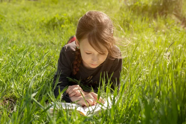 Маленька дівчинка лежить на животі, читаючи на траві. Вона має вигляд насолоди на обличчі, і вона виглядає дуже розслабленою — стокове фото