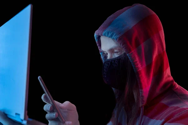 Ein männlicher Hacker im Sweatshirt mit Kapuze sitzt in einem Raum hinter einem Laptop und hält ein Telefon in der Hand. Cyberpunk-Stil — Stockbild Ein Männlicher Hacker Sweatshirt Mit Kapuze Sitzt Einem Raum Hinter — Stockfoto