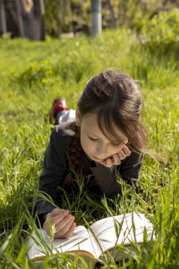 Küçük bir kız çimlerin üzerinde yatıp kitap okuyor. Yüzünde bir zevk ifadesi var ve çok rahat görünüyor..